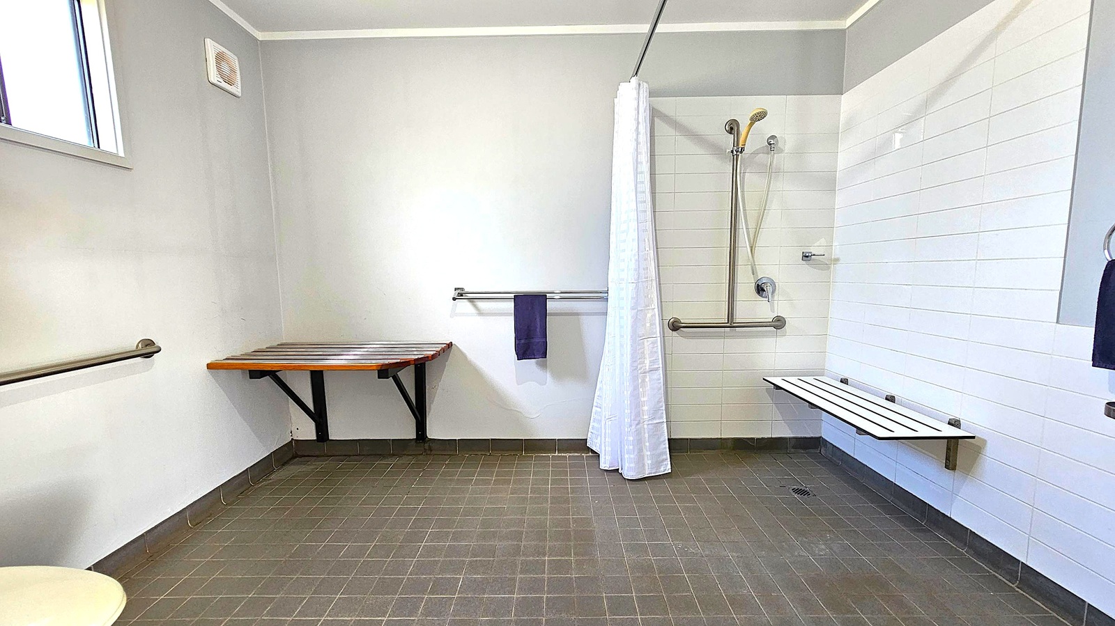 A bathroom with grey tiled flooring and light grey walls. On the left, a small window and a wooden bench are mounted on the wall. A metal grab bar runs along the wall. In the centre, a towel hangs on a rail beneath a white shower curtain. On the right, a shower area with a handheld showerhead and a horizontal grab bar. A white slatted bench is attached to the wall. The ceiling is plain and light-coloured.