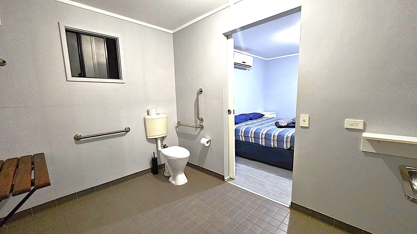 A bathroom with grey walls and a tiled floor. On the left, there's a small window and a wooden bench. In the centre, a toilet with a cistern is equipped with grab bars and a toilet roll holder. The door on the right leads to a bedroom with a bed covered in a striped duvet. An air conditioning unit is visible above the bed. The ceiling is plain white, and there are light switches on the right wall.