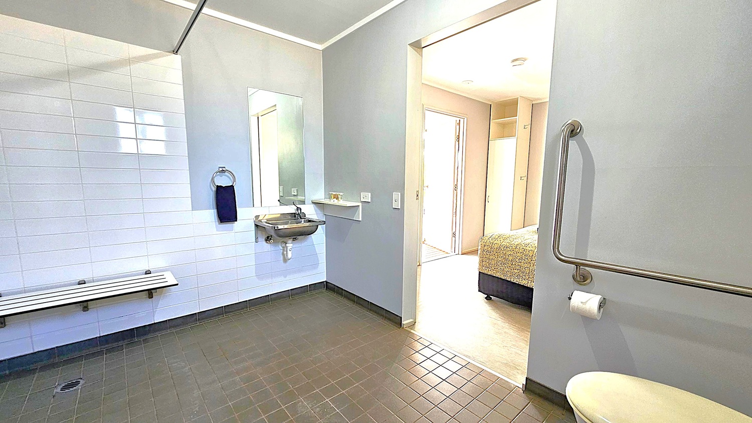 A bathroom with grey walls and dark floor tiles. On the left, there's a white-tiled wall with a foldable bench and a towel ring holding a dark towel. A small sink is mounted below a rectangular mirror. On the right, a toilet with a grab bar and toilet paper holder. The room opens into a bedroom with a bed visible, and a doorway leading to a well-lit area. The ceiling is plain and light-coloured.