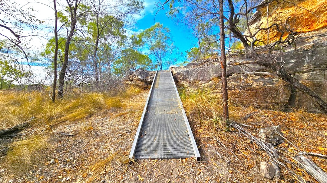 A narrow metal ramp leads upwards through a rocky landscape. On the left, sparse trees with thin trunks and dry grass are visible. On the right, a rocky wall with some overhanging branches and more dry vegetation. The ground is covered with dry leaves and twigs. In the background, more trees are visible against a blue sky.