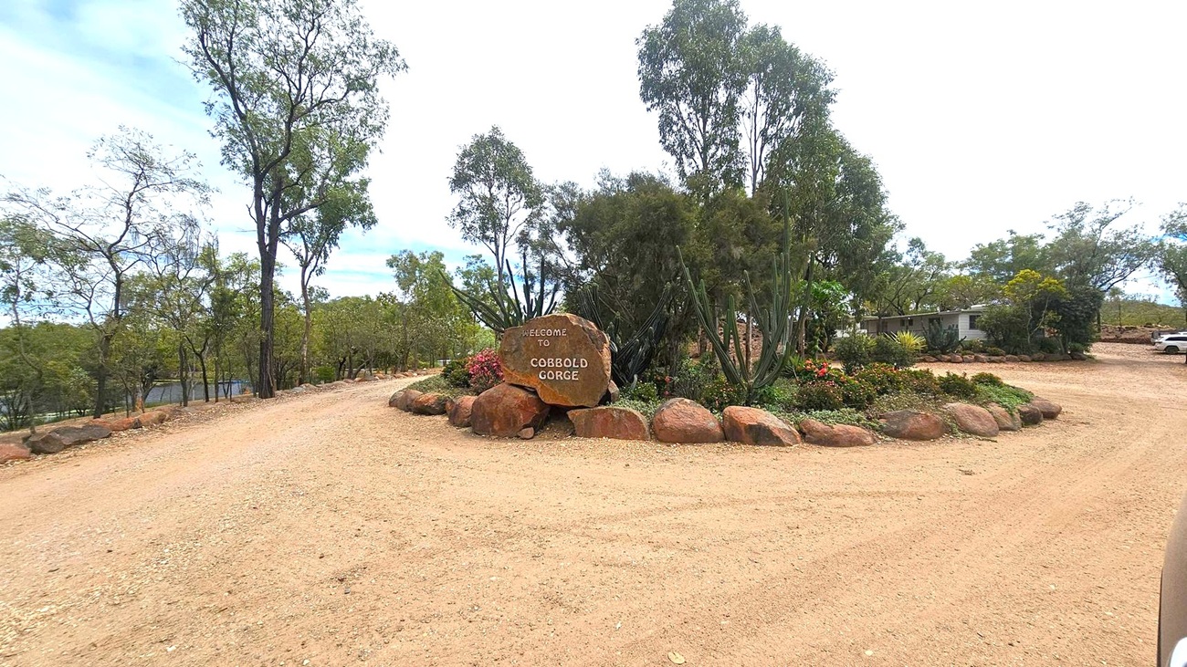A dirt path leads to a large rock with the words "Welcome to Cobbold Gorge" engraved on it, surrounded by smaller rocks. Trees and shrubs are scattered around, with a few flowering plants near the sign. To the right, a building is partially visible, and a white vehicle is parked nearby. The area is open with more trees in the background.