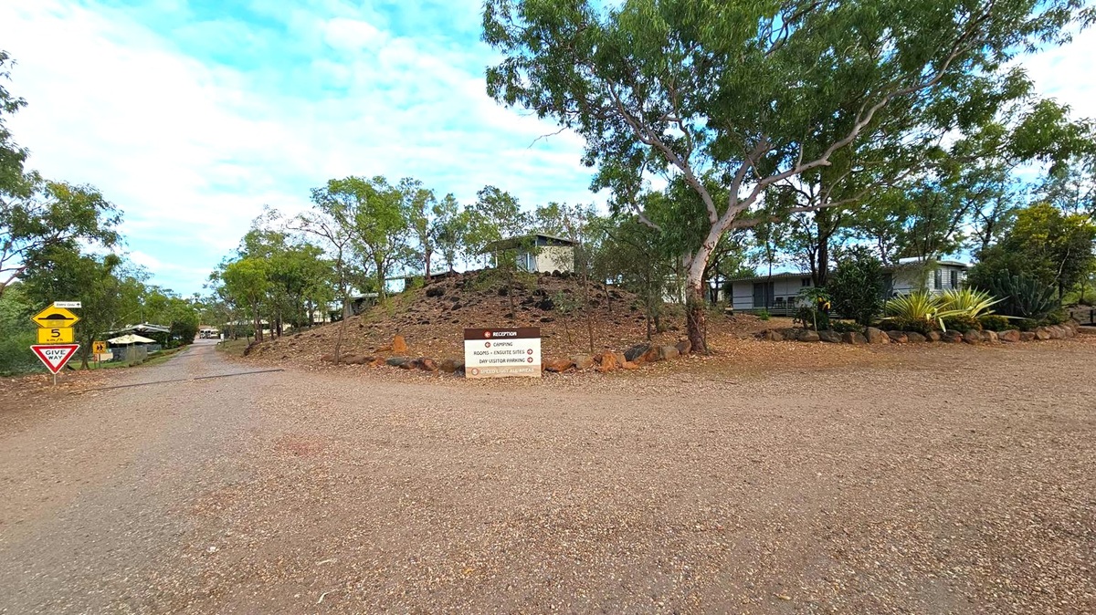 A gravel road leads into a rural area with trees and small buildings. On the left, there are traffic signs indicating a speed limit of 5 km/h and a "Give Way" sign. In the centre, a sign provides directions to reception, camping, rooms, ensuite sites, and day visitor parking. The right side features a rocky mound with scattered trees and a building partially visible behind it. The area is surrounded by greenery and a clear sky above.