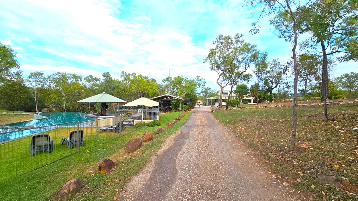 A gravel path runs through the centre, flanked by grass and scattered rocks. On the left, there's a fenced area with a swimming pool, sun loungers, and umbrellas. Trees surround the pool area, providing shade. On the right, more trees and a grassy area with patches of bare earth. In the background, there are buildings partially obscured by trees, and a person walking along the path. The sky is mostly clear with some clouds.
