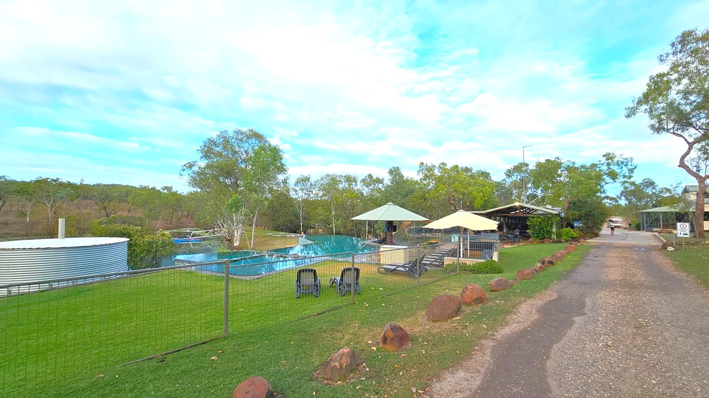 A grassy area with a metal fence on the left, enclosing a pool with sun loungers and two large umbrellas. A round metal water tank is visible near the pool. Trees surround the area, providing a natural backdrop. On the right, a gravel path leads to a building with a covered outdoor seating area. A person walks along the path, and a vehicle is parked further down. The scene is calm and open, with scattered rocks lining the path.