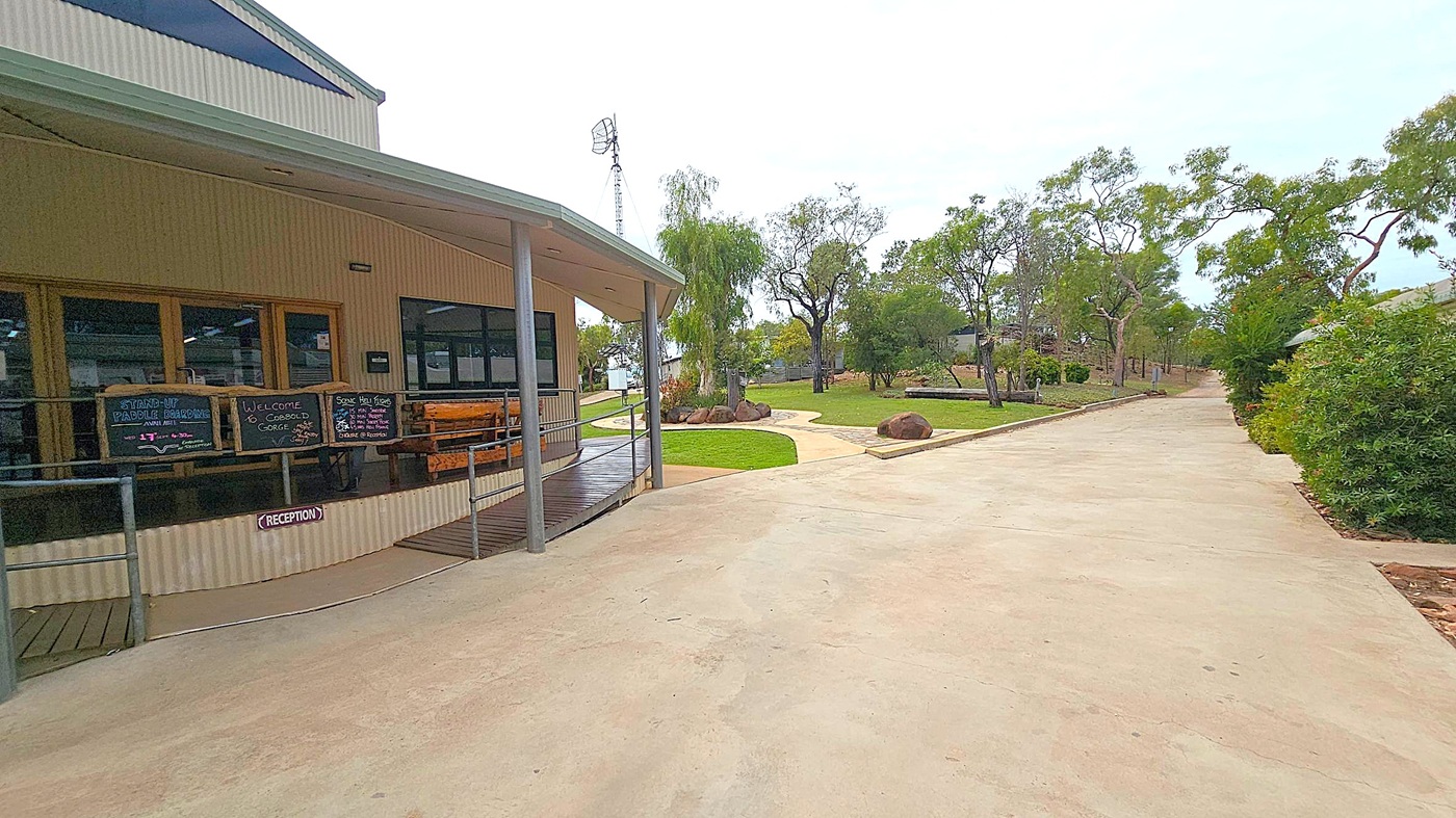 A building with a covered porch is on the left, featuring signs for activities like stand-up paddleboarding. A ramp leads to the entrance marked "Reception." On the right, a paved path extends into the distance, bordered by green grass and trees. Large rocks are scattered around the landscaped area. The background includes more trees and a tall antenna. The ground is a mix of concrete and grass, and the building has a corrugated metal exterior.