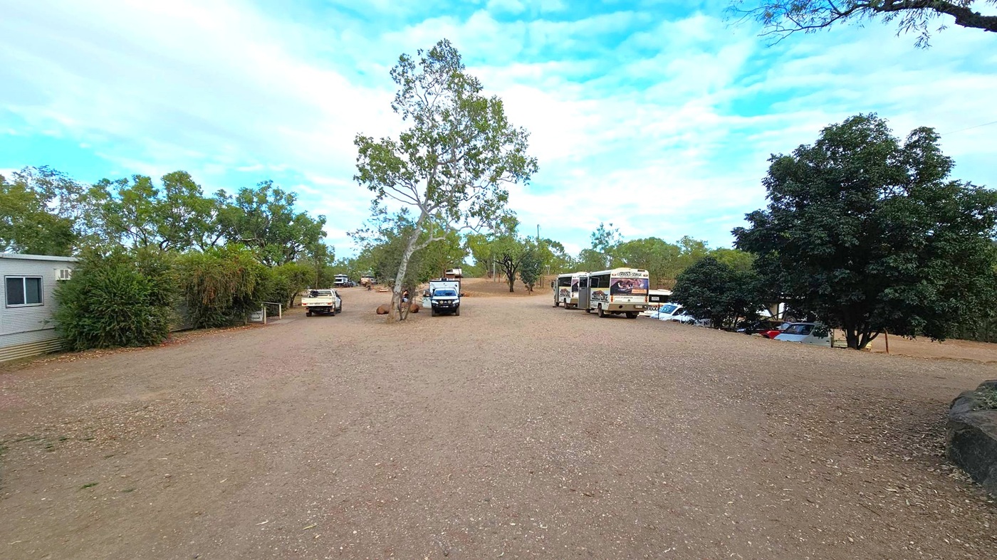 A dirt clearing with several vehicles parked, including a bus on the right. Trees are scattered throughout, with a prominent tree in the centre. On the left, a building with a window is partially visible, alongside some bushes. More vehicles and trees are in the background, creating a sparse, open area. The ground is covered in dirt and small stones.