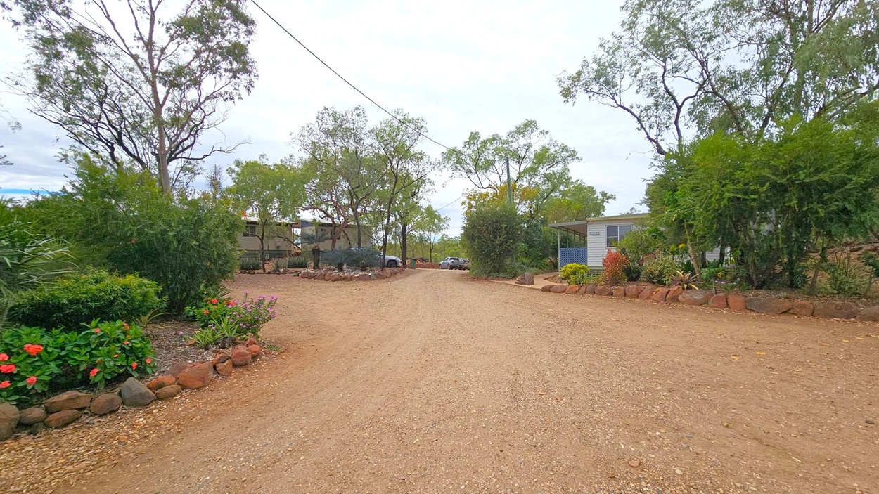 A gravel path runs through the centre, flanked by trees and shrubs. On the left, there are vibrant flowers and a small garden bordered by rocks. On the right, a building with a blue lattice porch is partially visible, surrounded by more greenery. In the background, additional structures and parked vehicles can be seen among the trees. The ground is a mix of gravel and dirt, and the area is enclosed by natural landscaping.