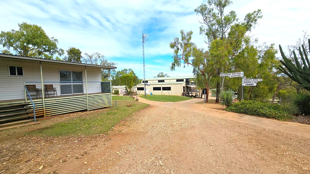 A gravel path leads through a rural area. On the left, a small building with a porch and two chairs is visible. In the centre, a larger building with a metal tower behind it is surrounded by trees. On the right, there are directional signs pointing to various facilities, and a large plant with spiky leaves. The ground is a mix of grass and dirt, and the area is dotted with trees.