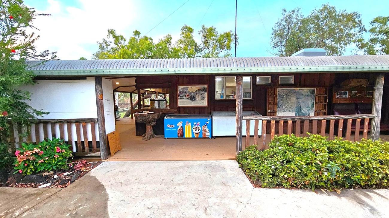 A small building with a corrugated metal roof and wooden posts. In the centre, there is an ice cream freezer with colourful advertisements. Inside, a map and some framed pictures are visible on the walls. The entrance is open, leading to a wooden floor. On the left, there are flowering plants and a small bush. On the right, a wooden railing with more greenery in front. Trees are visible in the background. The ground is a mix of concrete and soil.