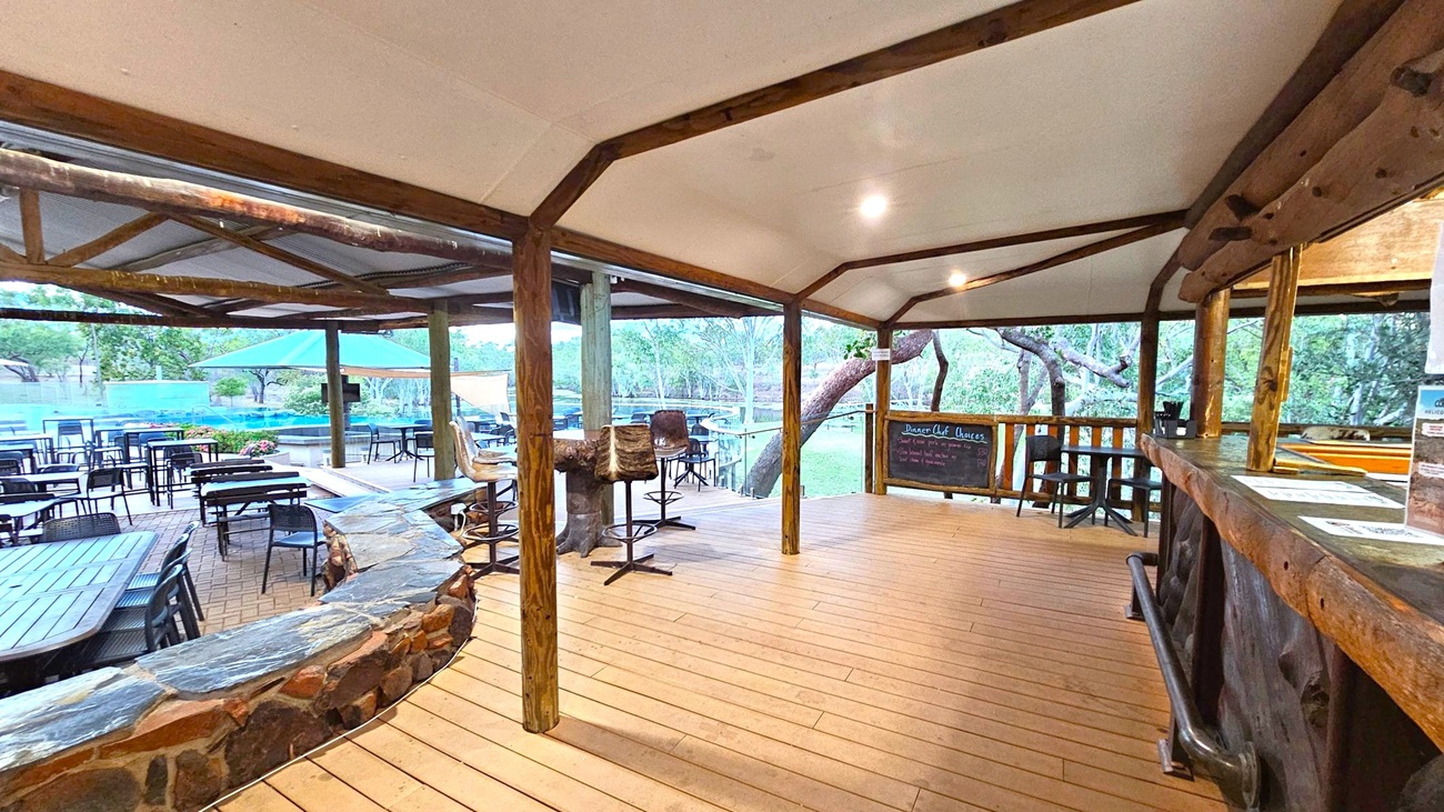 A rustic outdoor dining area with wooden flooring and a wooden ceiling supported by timber beams. On the right, there's a bar counter with menus and a chalkboard listing dessert options. In the middle, high stools with fur covers are placed around a table. On the left, several tables and chairs are arranged on a brick patio, with a stone wall separating the spaces. In the background, a large umbrella provides shade, and trees are visible beyond the patio.