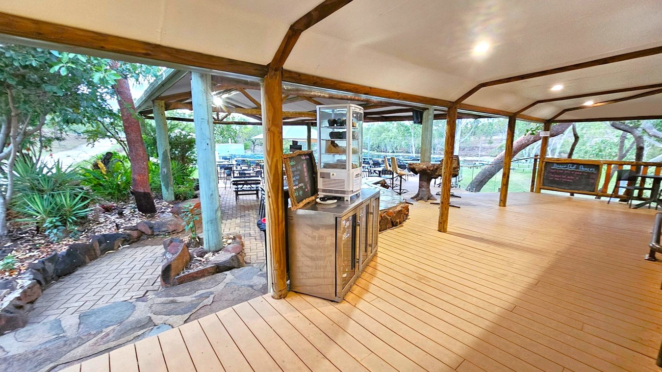 A covered outdoor area with a wooden floor and ceiling supported by wooden beams. In the centre, there's a metal counter with a glass display case and a chalkboard menu. To the left, a stone pathway leads through a garden with various plants and trees. In the background, there are tables and chairs set up on a brick patio, surrounded by more trees and greenery. A blackboard with writing is visible on the right side.