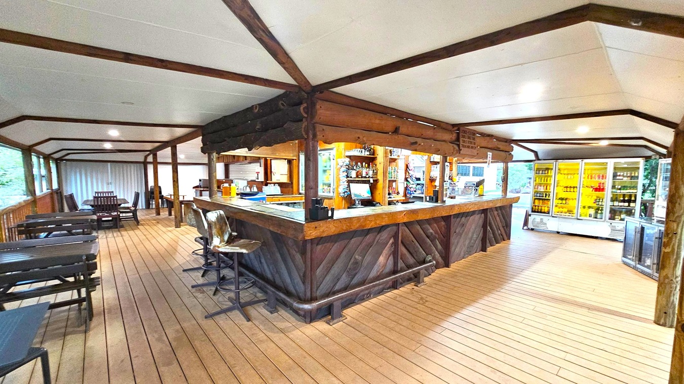 A rustic bar area with a wooden counter in the centre, surrounded by high stools. Behind the bar, shelves are stocked with various bottles and snacks. To the right, there are large refrigerators filled with drinks. On the left, several tables and chairs are arranged along the wooden floor. The ceiling is white with wooden beams, and the space is well-lit with recessed lighting. The background shows a view of greenery through large windows.