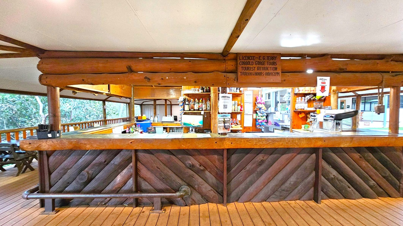 A rustic bar with a wooden counter and diagonal wood paneling. Behind the bar, shelves hold various bottles and snacks. A sign with trading hours is visible above. The ceiling is white, supported by wooden beams. The floor is made of wooden planks. To the left, there are outdoor seating areas with wooden tables and chairs, surrounded by greenery. The right side shows more shelves and a cash register.