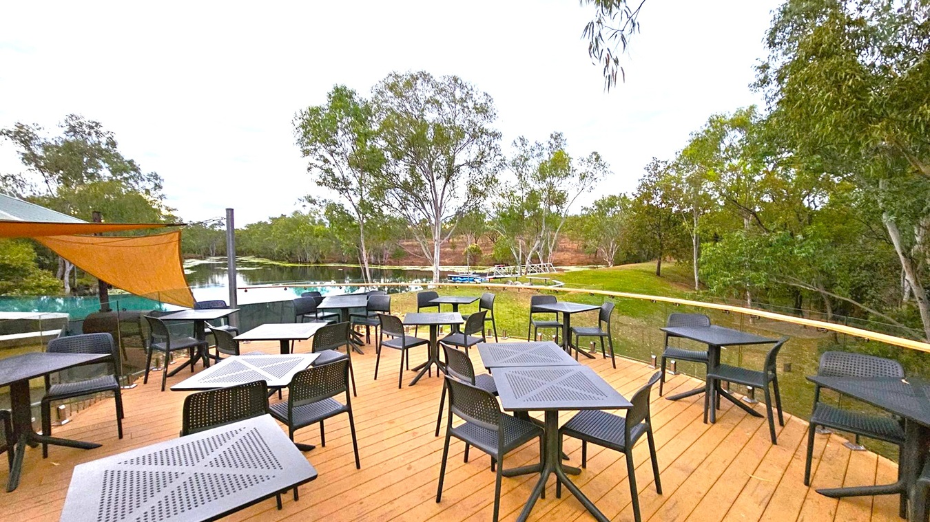 Outdoor seating area with several black tables and chairs on a wooden deck. To the left, a triangular shade sail is partially visible. In the background, there is a body of water surrounded by lush green trees. The area is enclosed with a glass railing, and the ground beyond the deck is grassy.