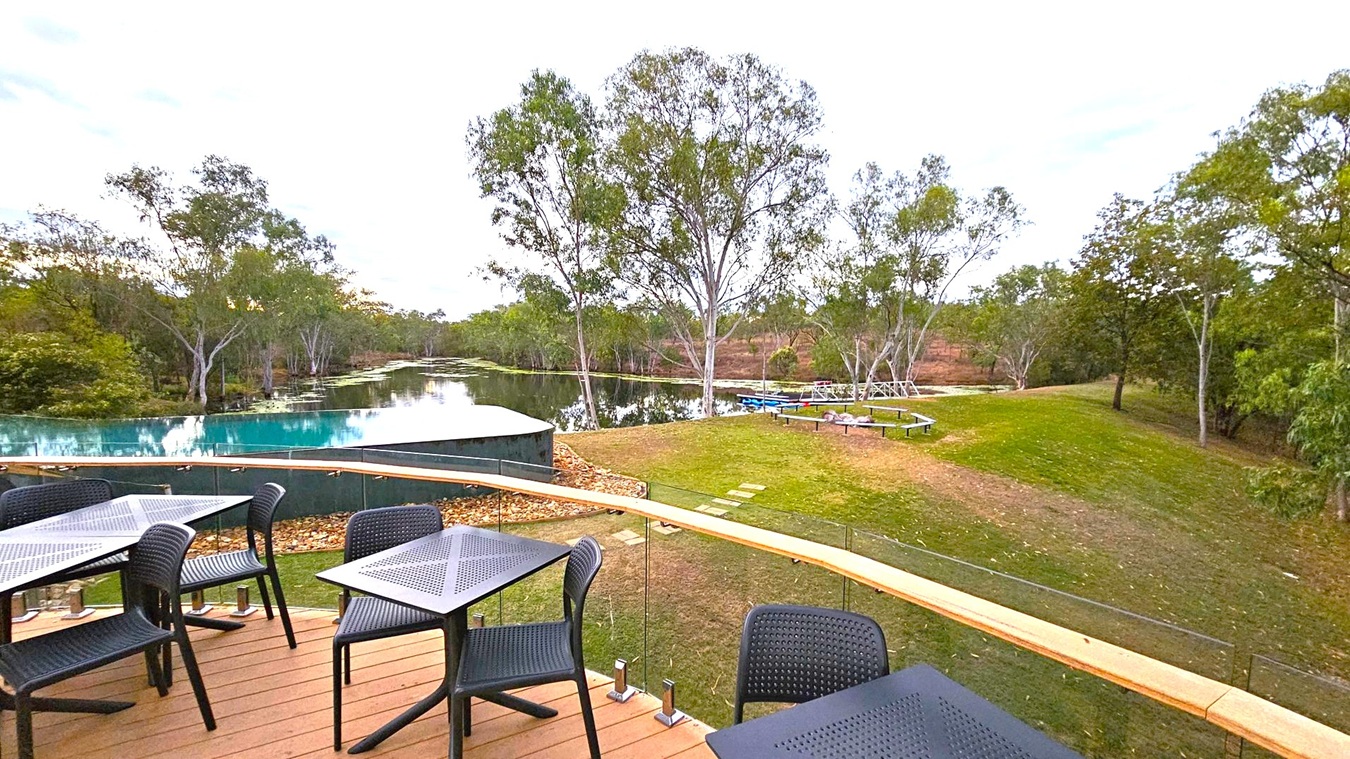 A wooden deck with black tables and chairs on the left overlooks an infinity pool that seamlessly blends into a natural body of water. The pool is bordered by a glass railing. In the background, tall trees with green foliage line the water's edge, and a grassy area with a few benches is visible on the right. The scene is tranquil, with a mix of natural and man-made elements.