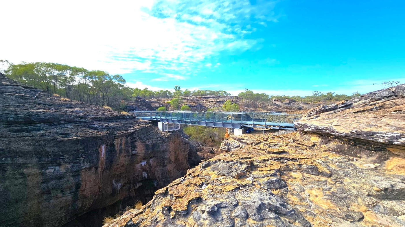 A rugged landscape with a metal bridge spanning a deep gorge. The foreground features rocky terrain with layers of exposed stone. The bridge, with railings, crosses from left to right, connecting the rocky outcrops. In the background, sparse trees and shrubs are scattered across the rocky hills. The sky is mostly clear with some clouds.