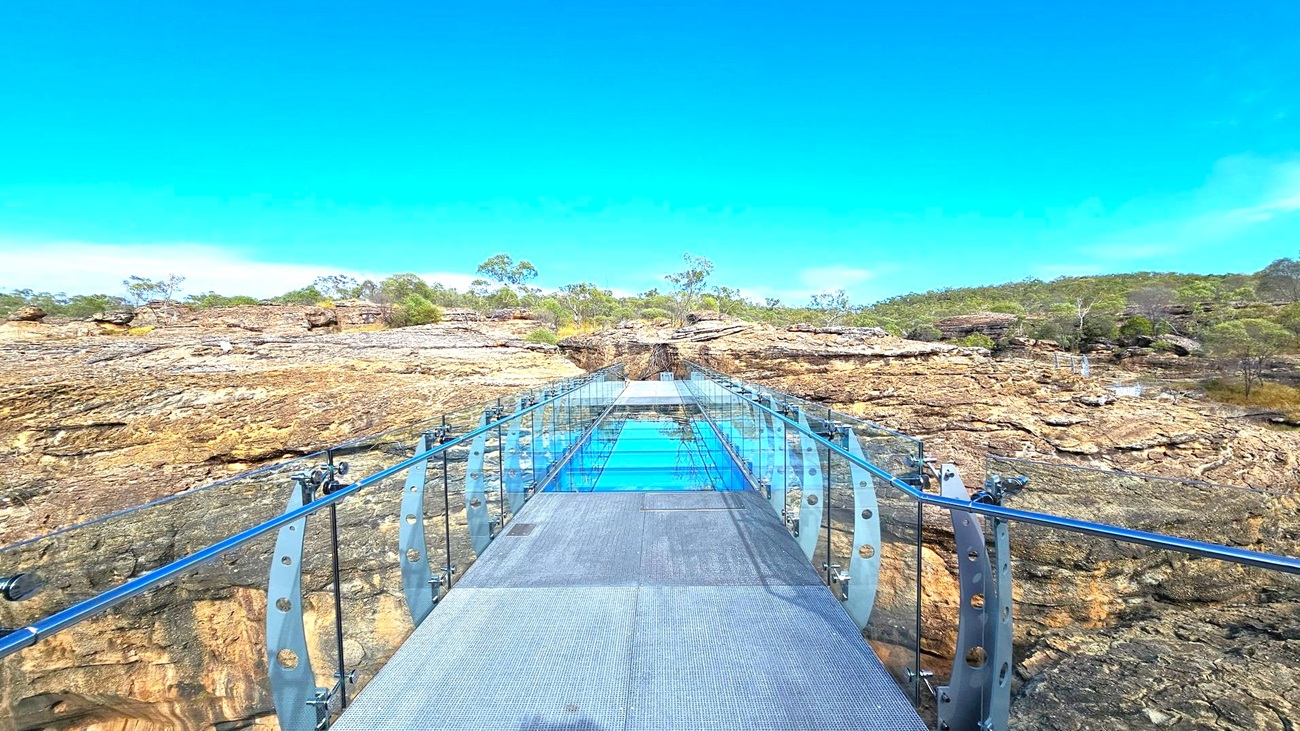 A glass-bottomed bridge extends across a rocky landscape. The bridge has transparent panels and metal railings on both sides. The rocky terrain below is rugged, with patches of greenery and sparse trees. In the background, more rocks and trees are visible, creating a natural setting. The sky is clear and blue, providing a bright backdrop.