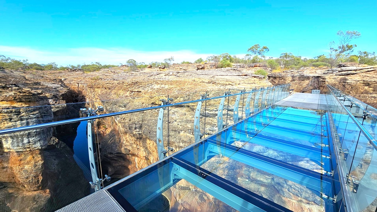 A glass-bottomed bridge extends over a deep gorge, with clear panels and metal railings on both sides. The bridge reflects the blue sky above. The surrounding landscape features rugged, rocky terrain with sparse vegetation and small trees scattered across the area. The gorge below is steep and shadowed, contrasting with the bright, open space above.