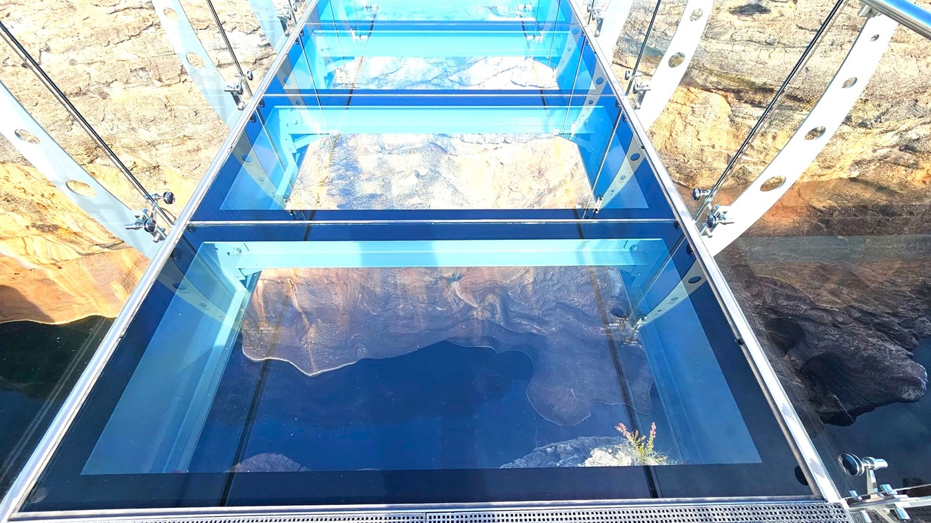 A glass-bottomed bridge with metal supports and cables spans across a rocky canyon. The transparent floor reveals a view of the canyon below, with rugged rock formations visible through the glass. The bridge is supported by metal beams and cables, with circular cutouts in the side panels. The surrounding landscape consists of textured rock surfaces with varying shades of brown and grey.