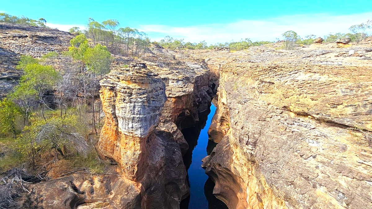 A narrow gorge with steep, rocky walls stretches through the centre, with a small stream of water visible at the bottom. The rock formations are layered and rugged, displaying shades of brown and grey. Sparse vegetation, including small trees and shrubs, grows on the rocky terrain on both sides of the gorge. The landscape is arid, with a clear blue sky above.