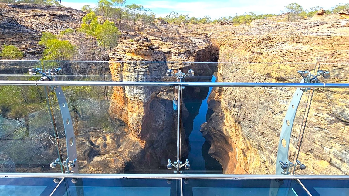 A glass-bottomed viewing platform with metal railings overlooks a deep, narrow gorge with steep, rocky walls. The gorge has a small body of water at the bottom, reflecting the surrounding rock formations. Sparse vegetation, including small trees and shrubs, grows on the rocky terrain. The background features a rugged landscape with more rocky outcrops and scattered greenery.