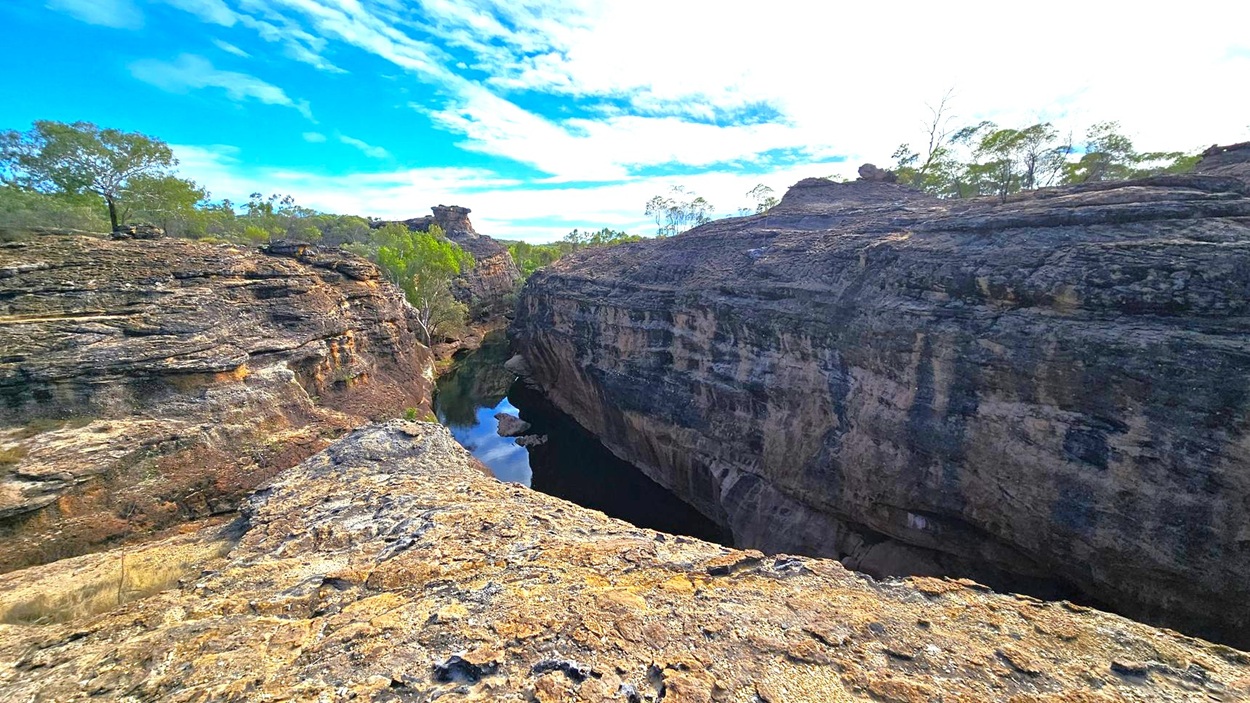 Rocky landscape with a deep gorge running through the centre. The foreground features rugged, layered rock formations. On the left, sparse trees and shrubs grow along the edge of the gorge. The right side shows more rock formations with some vegetation on top. In the middle, a narrow water body reflects the rocks above. The background has more trees and rock formations under a partly cloudy sky.