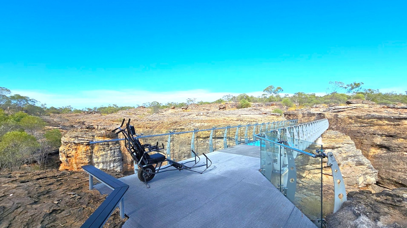 A glass-floored bridge extends across a rocky landscape. On the left, a black wheelchair is positioned on the bridge's platform, which has a metal railing. The bridge is supported by glass panels with metal brackets. In the background, there are rugged rock formations and sparse vegetation, including small trees and shrubs. The ground is uneven with exposed rock surfaces.