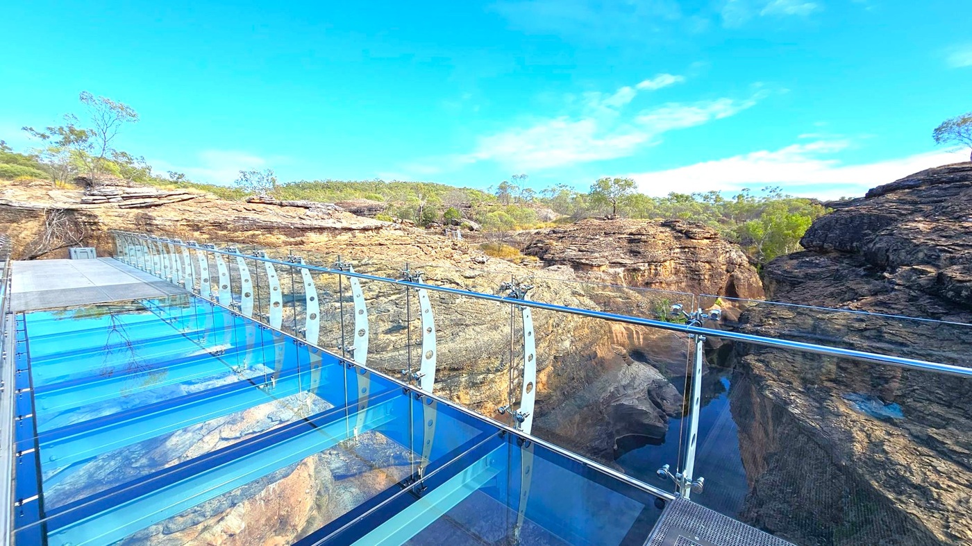 A glass-bottomed walkway extends over a rocky canyon, with transparent panels and metal railings. The left side shows the walkway leading to a rocky terrain with sparse vegetation. On the right, the canyon drops steeply, revealing rugged rock formations and a narrow waterway below. In the background, a landscape of rocky outcrops and scattered trees stretches into the distance. The sky is clear and blue.
