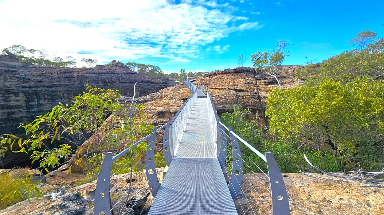 A narrow metal bridge with railings stretches across a rocky landscape. On the left, sparse vegetation and rocky formations are visible, while on the right, dense green foliage and trees are present. The bridge leads towards a rugged terrain with more rocks and scattered trees in the background. The sky is partly cloudy with patches of blue.