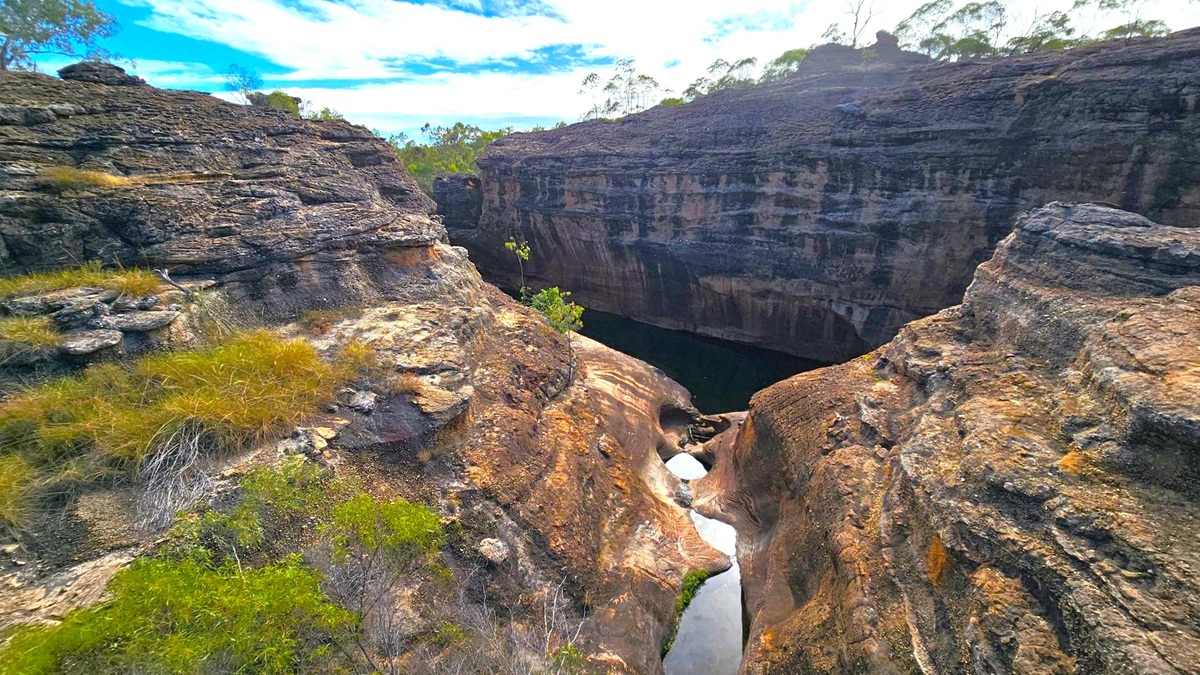 Rocky landscape with a deep gorge in the centre. The foreground features rugged, layered rock formations with patches of dry grass and small shrubs. A narrow stream of water runs through the middle, disappearing into the shadowy depths of the gorge. The background shows more rock formations and sparse vegetation, with trees silhouetted against the sky.