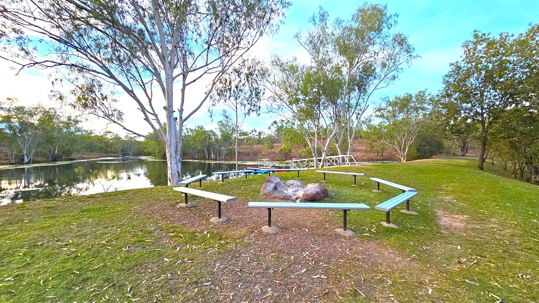 A serene park setting with a circular arrangement of benches surrounding a cluster of large rocks in the centre. The ground is covered with grass and scattered leaves. In the background, a calm lake reflects the surrounding trees, which are tall and sparse with slender trunks. A small dock extends into the water. The sky is partly cloudy, and the area is surrounded by lush greenery.