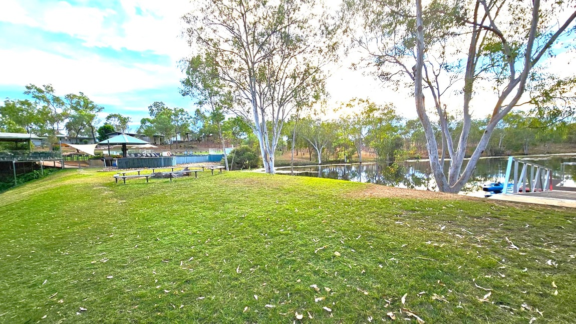 Grassy area with scattered leaves in the foreground. On the left, there are several benches and a shaded area with a canopy, adjacent to a wooden deck. In the centre, tall trees with slender trunks and sparse foliage. On the right, a small dock extends into a calm body of water, with a blue kayak visible. The background features more trees and a few structures, possibly buildings, partially obscured by the foliage.