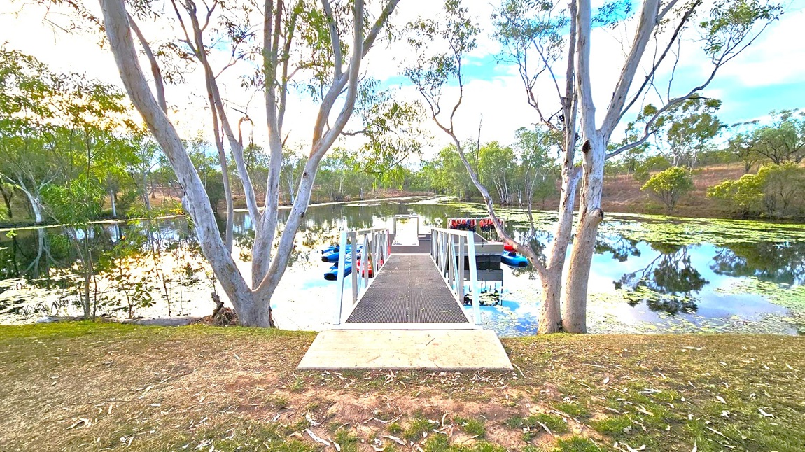 A narrow metal walkway extends from the foreground to a floating dock on a calm lake. The dock is surrounded by several kayaks and paddle boats. Tall trees with slender trunks and sparse foliage frame the scene on both sides. The water reflects the trees and sky, with patches of green lily pads scattered across the surface. The ground in the foreground is a mix of grass and bare earth, with some fallen leaves.