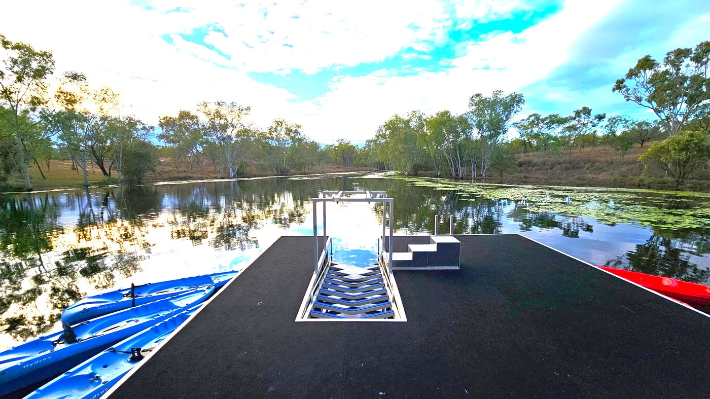 A tranquil lake scene with a black platform in the foreground, featuring a metal ladder and steps leading into the water. On the left, several blue kayaks are visible. The background is filled with trees lining the water's edge, and the surface of the lake reflects the sky and surrounding greenery.