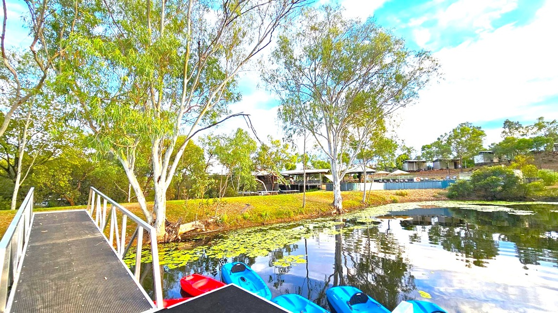 A metal walkway extends over a body of water in the foreground, leading to a grassy bank. Several kayaks, red and blue, are moored at the water's edge. The water is dotted with lily pads. Tall trees with slender trunks and green foliage line the bank. In the background, there are structures resembling cabins or shelters, partially obscured by trees. The sky is partly cloudy, with patches of blue visible.