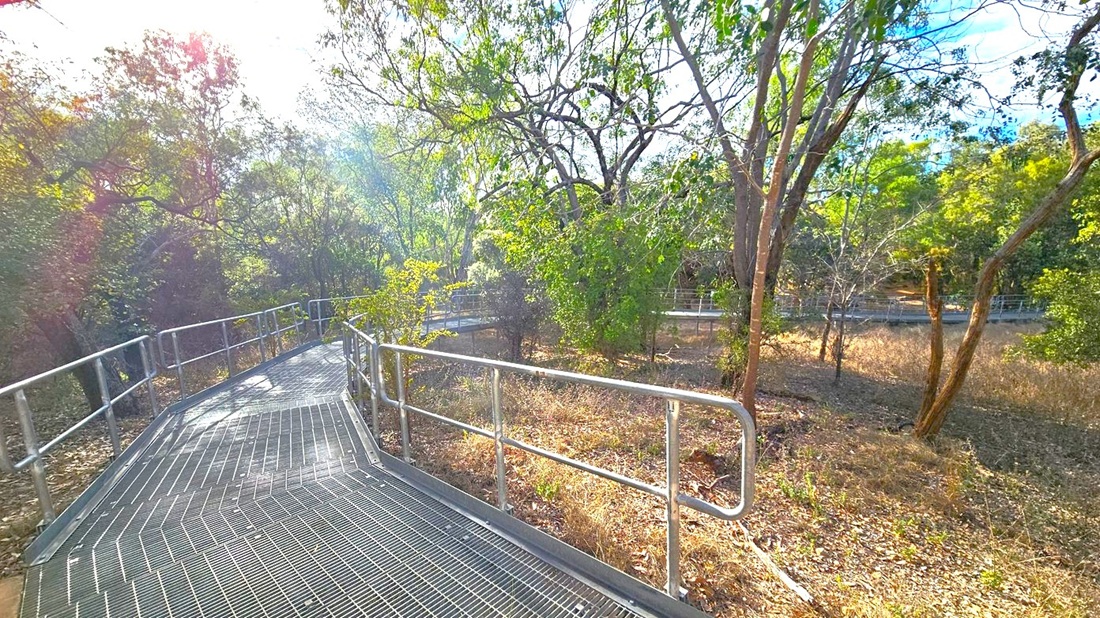 A metal walkway with railings curves through a wooded area. On the left, dense trees with green foliage partially obscure the view. On the right, more trees and dry grass cover the ground. The sun is visible through the trees, casting light and creating lens flares. The walkway is elevated, with a grated surface, and the surrounding area is natural and untamed.