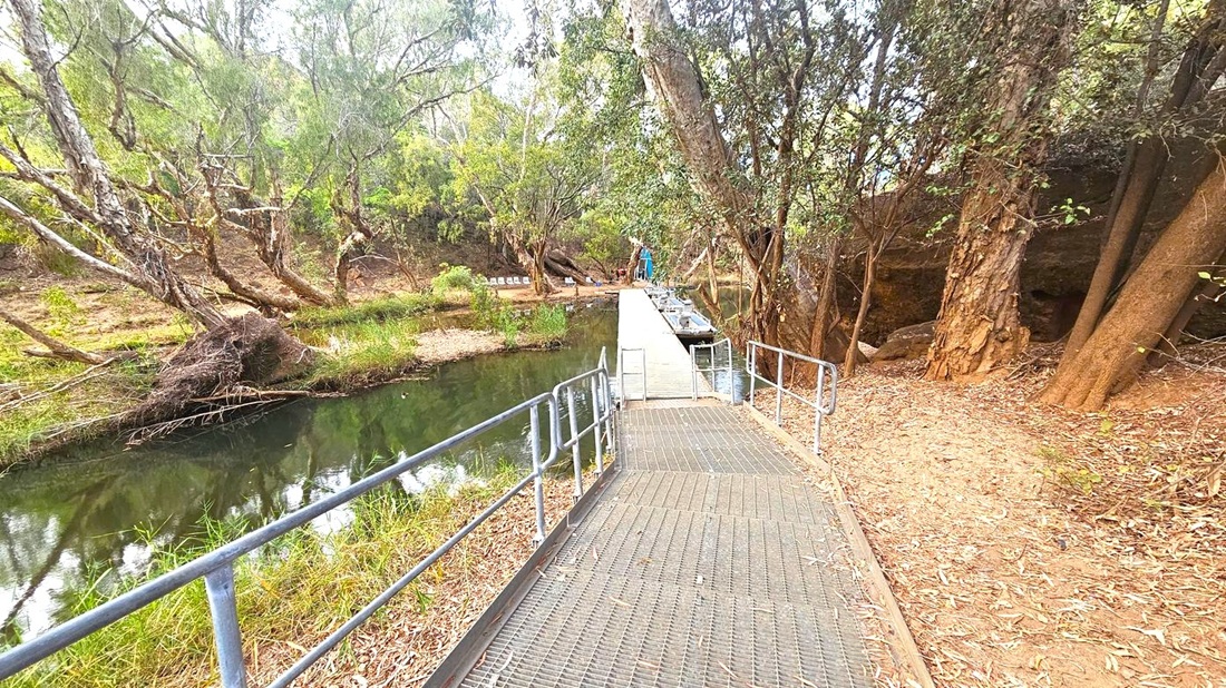A narrow metal walkway with railings extends over a calm, narrow river. On the left, the riverbank is lined with dense trees and shrubs, their branches extending over the water. On the right, the ground is covered with dry leaves and more trees, with a dirt path running alongside. In the background, the walkway leads to a small dock area with boats moored and a few chairs visible. The scene is surrounded by a natural, wooded environment.