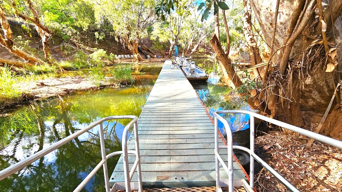 A narrow metal walkway extends over a calm body of water, flanked by railings. On the right, a small boat is moored, and a bicycle is visible further along the walkway. The surrounding area is lush with tall trees and dense foliage, casting reflections on the water. The ground is covered with dry leaves and twigs. In the background, there are several chairs arranged on the bank, suggesting a resting area.