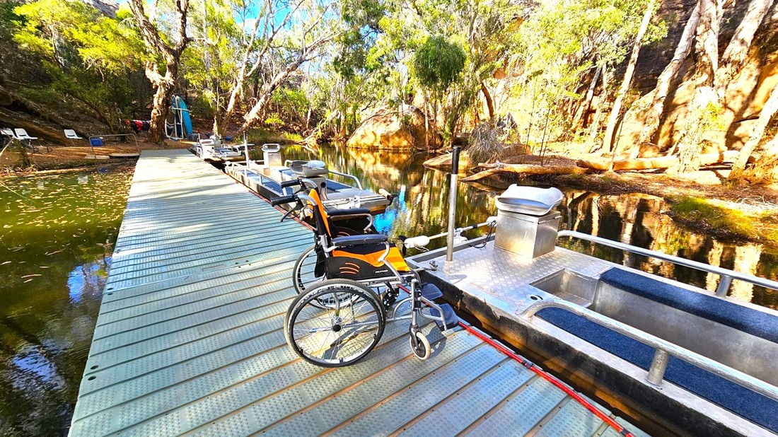 A metal dock extends over a calm body of water, with a wheelchair positioned on it. To the right, a small boat with metal railings and a seat is moored. The background features a rocky, tree-lined bank with dense foliage and a large boulder. The water reflects the surrounding trees and rocks. On the left, chairs and equipment are visible on the bank. The dock surface is textured with diagonal lines.