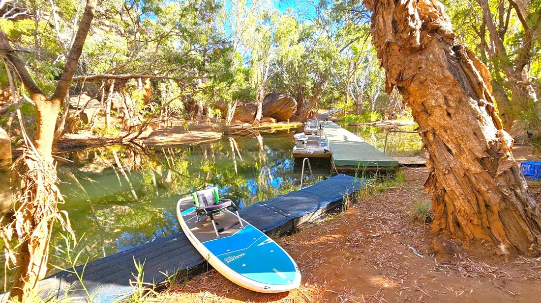 A serene river scene with a paddleboard resting on a small dock in the foreground. The river is calm, reflecting the surrounding trees and vegetation. A larger wooden dock extends into the water, equipped with several small outboard motors. The area is surrounded by dense trees with rough bark, and the ground is covered in dry leaves and grass. The setting is tranquil, with natural greenery and a clear view of the water.