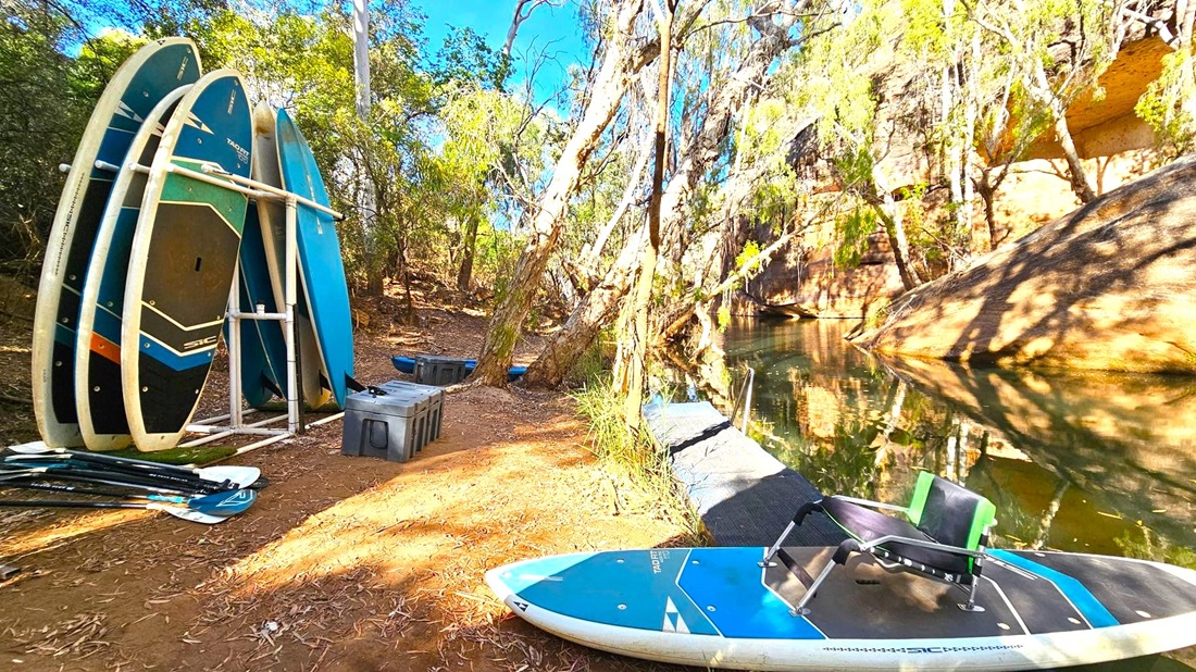 On the left, several paddleboards are stacked vertically on a white rack, with paddles lying on the ground nearby. In the centre, a dirt path is surrounded by trees with a few storage boxes scattered around. On the right, a paddleboard with a seat is positioned on a small dock leading to a calm body of water. The background features a rocky cliff face partially covered by trees, casting shadows on the water.