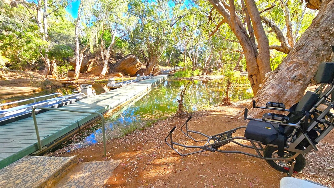 A serene natural setting with a wooden dock extending over a calm body of water on the left. The dock has railings and is surrounded by lush greenery and tall trees. On the right, a specialised wheelchair is positioned on a dirt path, leaning against a large tree. The ground is covered with dry leaves and the area is shaded by the tree canopy.