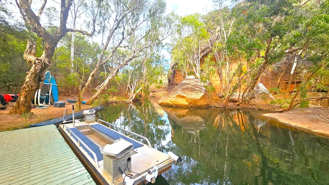 A serene waterway with a small boat docked on a wooden platform in the foreground. To the left, several paddleboards are propped against a tree, with more trees and some equipment nearby. The right side features a large rock formation partially submerged in the water, surrounded by trees. The background is filled with dense greenery and a rocky cliff, with reflections visible on the calm water surface.