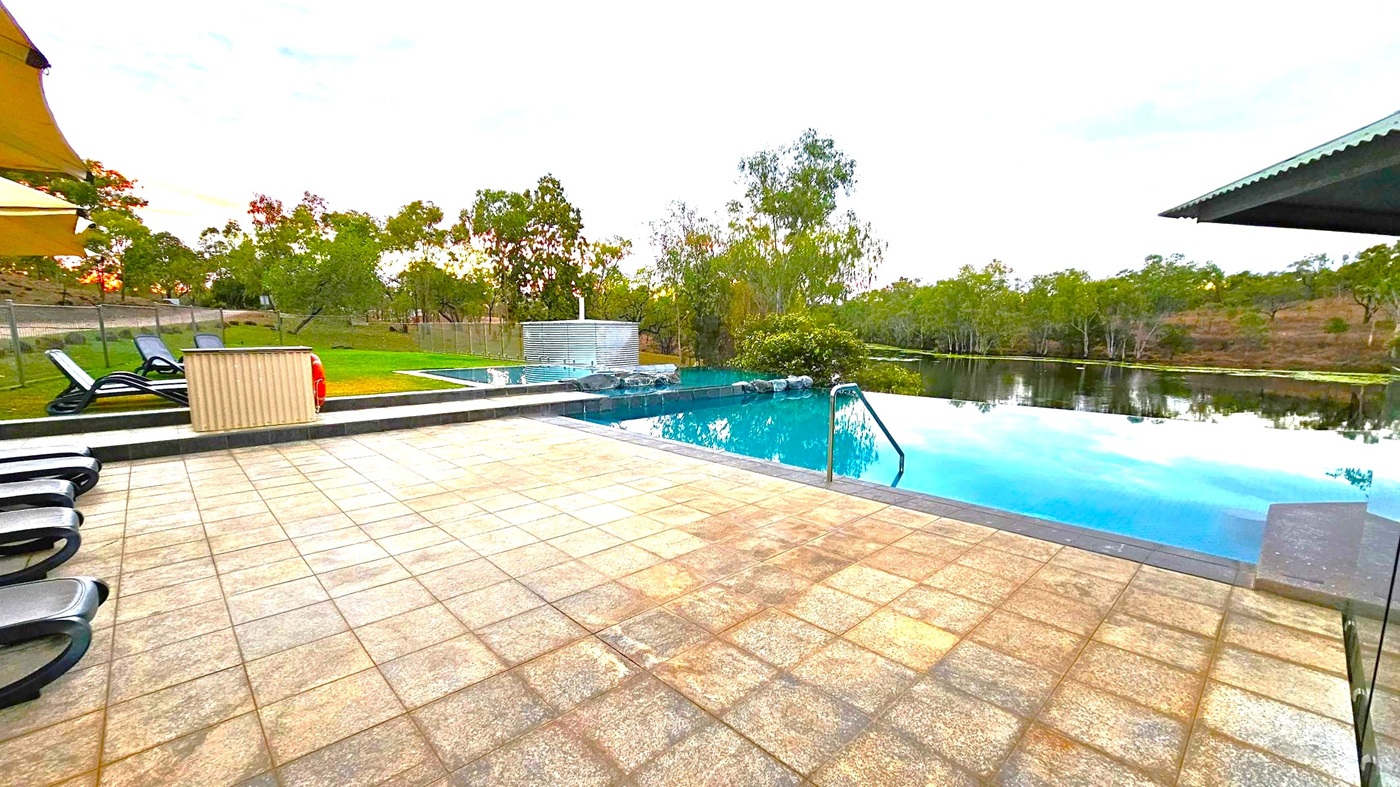 A serene outdoor setting with an infinity pool on the right, reflecting the surrounding trees and sky. The pool is bordered by a tiled patio area with several black sun loungers on the left. In the background, there are trees and a grassy area, with a fence running along the perimeter. A small structure with a metallic finish is near the pool. The sky has a mix of clouds with hints of pink and orange.