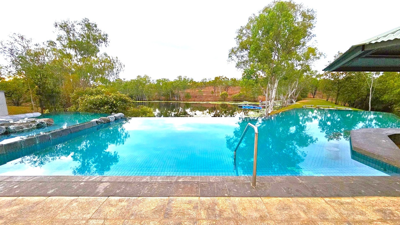 An infinity pool with clear blue water is in the foreground, bordered by a stone deck. A metal handrail descends into the pool. In the background, there are lush green trees and a calm body of water reflecting the sky. To the right, a structure with a corrugated metal roof is partially visible. The scene is serene, with natural greenery surrounding the pool area.
