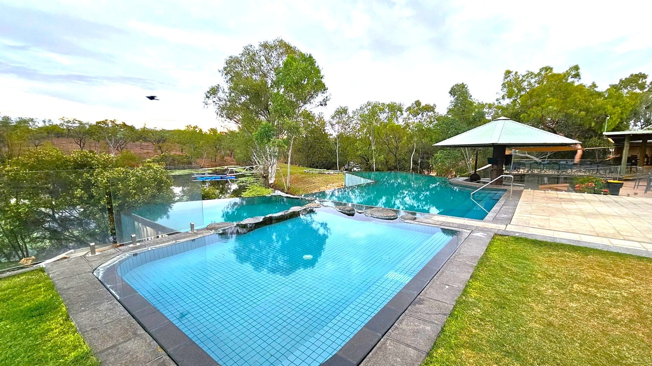 A serene outdoor setting with two connected infinity pools. The foreground features a smaller pool with a tiled floor, bordered by stone. To the right, a larger pool extends towards a gazebo with a green roof, surrounded by a paved area. Lush greenery and trees fill the background, with a glimpse of a river or lake beyond the pools. A bird is captured mid-flight in the sky.
