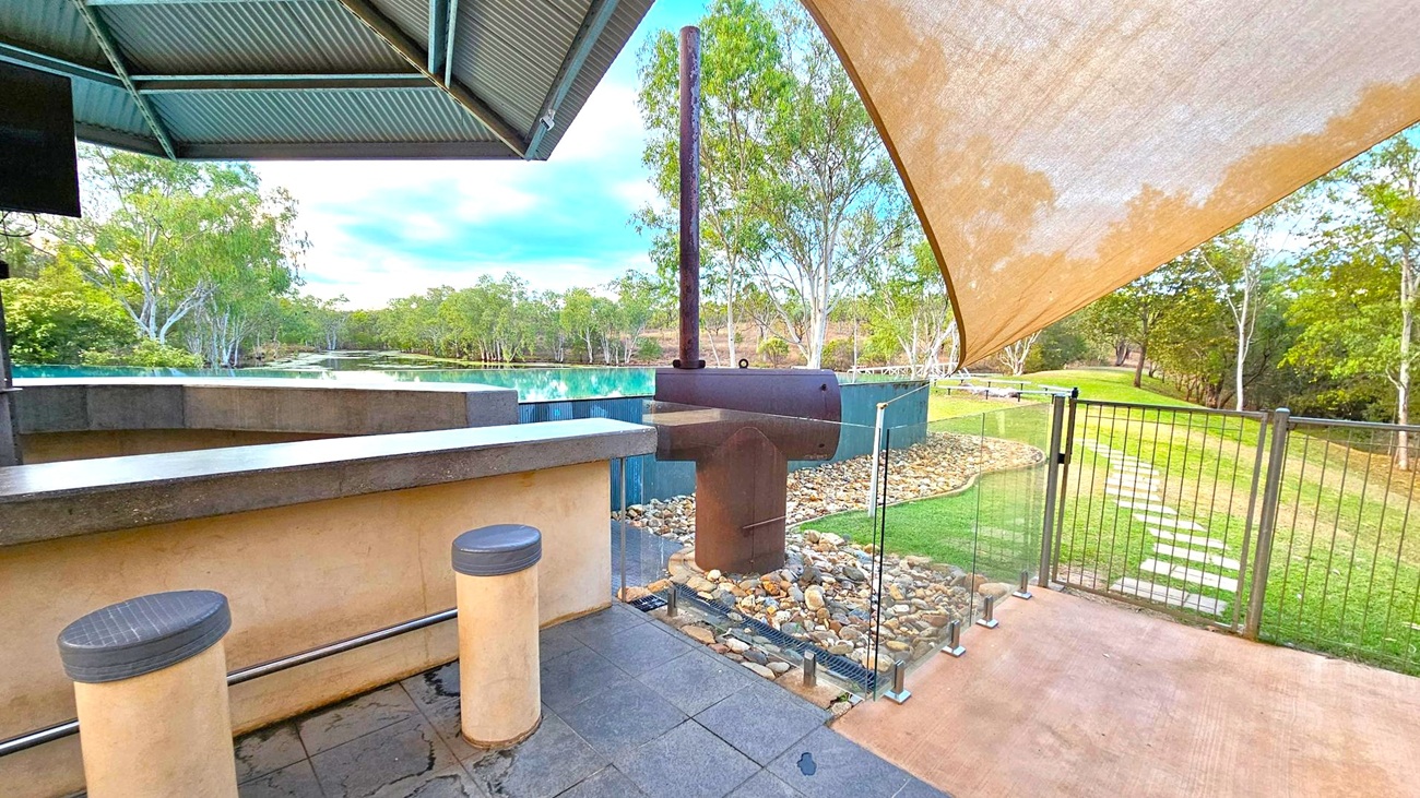A covered outdoor area with a metal roof and a bar counter with two round stools on the left. In the centre, there's a barbecue grill next to a glass fence. The ground is paved with stone tiles and pebbles, leading to a grassy area with stepping stones. In the background, a body of water is surrounded by trees, and a large shade sail extends overhead on the right.