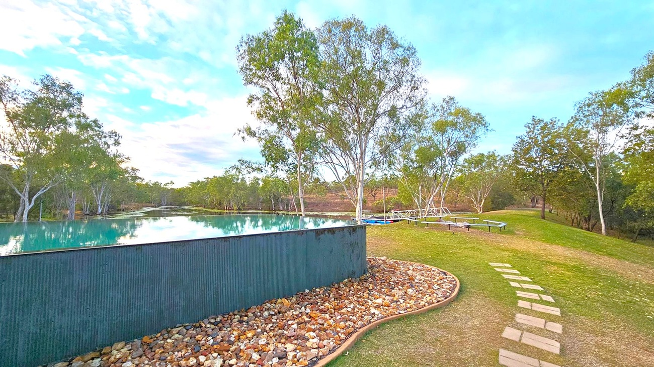 A serene landscape with an infinity pool on the left, bordered by a corrugated metal wall and a bed of small rocks. The pool reflects the surrounding trees and sky. On the right, a grassy area features a path made of stepping stones leading to picnic tables. Tall trees with slender trunks and green foliage are scattered throughout the scene, with more dense greenery in the background.