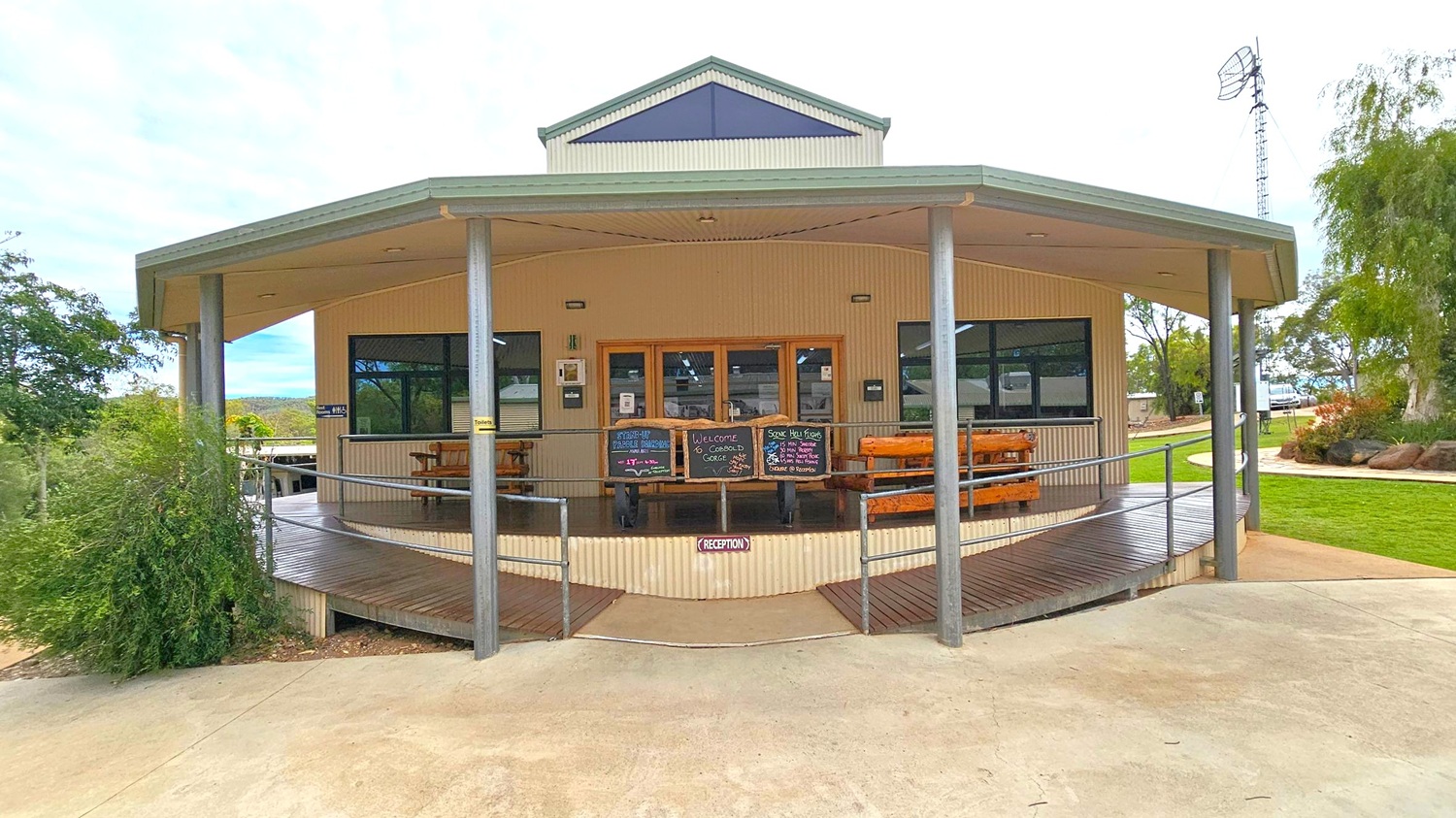 A single-storey building with a light-coloured exterior and a green-trimmed roof. The front features large windows and a central wooden door, flanked by chalkboards with writing. A wooden deck with picnic tables is supported by metal poles. A ramp leads up to the entrance, with a "Reception" sign at the base. Surrounding the building are green lawns and trees, with a tall antenna visible on the right. The ground is paved with concrete.