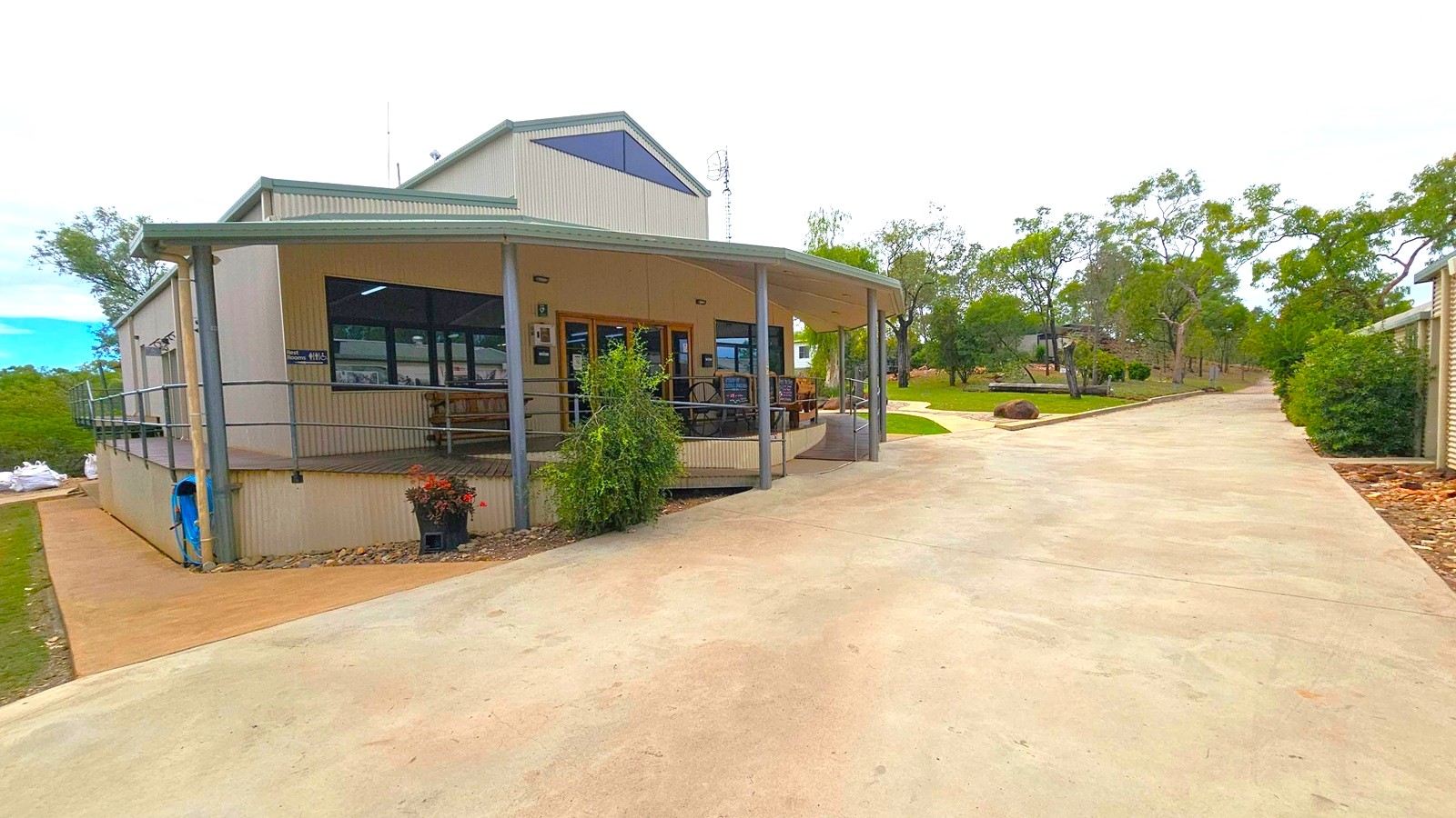 A single-storey building with a corrugated metal exterior and a sloped roof is on the left. It has a covered porch with railings and several windows. A concrete path leads up to the entrance, with potted plants near the doorway. On the right, a wide concrete driveway extends into the distance, bordered by trees and shrubs. The background features more trees and a grassy area, with a small structure visible further back.